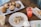A variety of pastries and desserts are arranged on plates atop a rustic wooden table. There are muffins with different toppings and flavors, cookies, a cannoli, and small macarons. Two beverages in plastic cups, one iced coffee and another with a pink hue and fruit slices, are also visible.