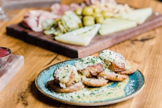 A plate of breaded and fried food items topped with grated cheese and herbs is in the foreground. The food is served on a turquoise plate placed on a wooden table. In the background, there is a wooden board with a variety of foods, including sliced meats, cheeses, and pickled items.