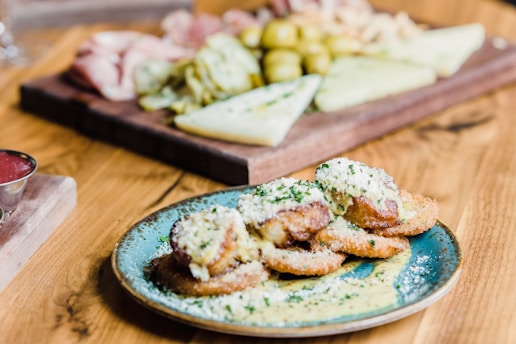 A plate of breaded and fried food items topped with grated cheese and herbs is in the foreground. The food is served on a turquoise plate placed on a wooden table. In the background, there is a wooden board with a variety of foods, including sliced meats, cheeses, and pickled items.