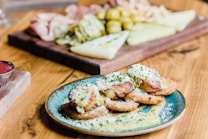 A plate of breaded and fried food items topped with grated cheese and herbs is in the foreground. The food is served on a turquoise plate placed on a wooden table. In the background, there is a wooden board with a variety of foods, including sliced meats, cheeses, and pickled items.
