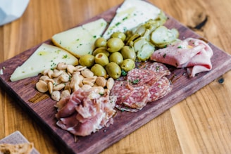 A wooden board is topped with a variety of charcuterie items, including sliced salami, prosciutto, and folded ham. Accompanying these meats are ripe green olives and slices of pickles. There are also several wedges of firm cheese and a pile of almonds. Everything is neatly arranged and sprinkled with herbs for garnish.