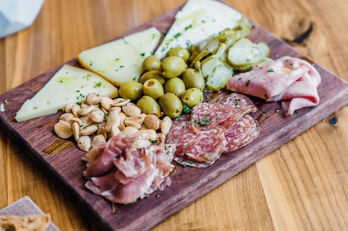 A wooden board is topped with a variety of charcuterie items, including sliced salami, prosciutto, and folded ham. Accompanying these meats are ripe green olives and slices of pickles. There are also several wedges of firm cheese and a pile of almonds. Everything is neatly arranged and sprinkled with herbs for garnish.