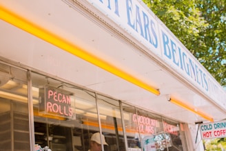A food stand with signs advertising pecan rolls and beignets. Neon lights display the words 'Pecan Rolls' and 'Chocolate Eclairs'. A person wearing a white cap can be seen inside the stand. The exterior is white with yellow accents and trees are visible in the background.