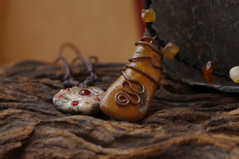 Two handcrafted necklaces are displayed on a textured wooden surface. The first pendant resembles a polished stone wrapped in copper wire, while the second one is a metallic disk adorned with red gems. A string of amber-like beads accompanies the first pendant, adding to the earthy aesthetic.