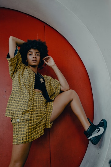 A person with curly hair poses confidently against a bold red circular background. They are dressed in a matching yellow and black patterned outfit, consisting of a jacket and shorts, paired with stylish high-top sneakers.