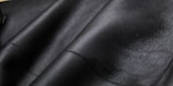Close-up texture of black leather material with visible grain and subtle shine. The surface appears smooth and slightly glossy, showcasing the quality of the material.