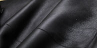 Close-up texture of black leather material with visible grain and subtle shine. The surface appears smooth and slightly glossy, showcasing the quality of the material.