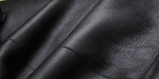 Close-up texture of black leather material with visible grain and subtle shine. The surface appears smooth and slightly glossy, showcasing the quality of the material.