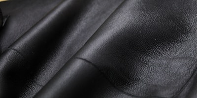 Close-up texture of black leather material with visible grain and subtle shine. The surface appears smooth and slightly glossy, showcasing the quality of the material.