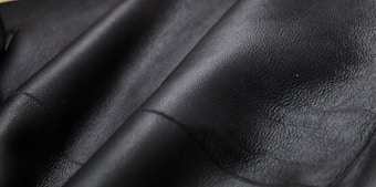Close-up texture of black leather material with visible grain and subtle shine. The surface appears smooth and slightly glossy, showcasing the quality of the material.