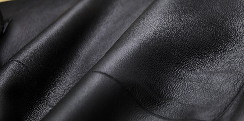 Close-up texture of black leather material with visible grain and subtle shine. The surface appears smooth and slightly glossy, showcasing the quality of the material.