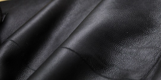 Close-up texture of black leather material with visible grain and subtle shine. The surface appears smooth and slightly glossy, showcasing the quality of the material.