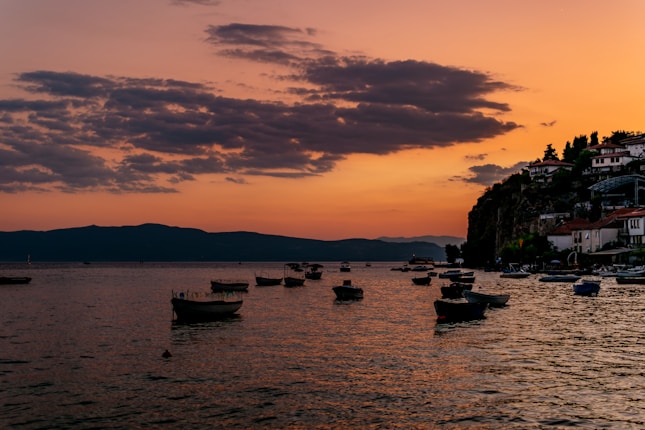 A serene lakeside scene with a number of small boats floating on the water. The sky is illuminated with vibrant hues of orange and purple as the sun sets, casting a warm glow over the scene. On the right, a cliff is topped with charming buildings and a few trees, adding to the picturesque quality of the landscape. The distant hills are faintly visible under the fading light.