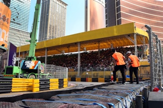 A busy urban scene featuring a large spectator stand filled with people. The stand is covered by a yellow canopy, and there is a green construction vehicle with a 'Macau Grand Prix' sign in front. Two individuals in orange vests are standing near the barriers that line a racetrack. Skyscrapers surround the area, creating a city backdrop.