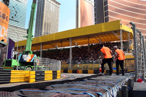 A busy urban scene featuring a large spectator stand filled with people. The stand is covered by a yellow canopy, and there is a green construction vehicle with a 'Macau Grand Prix' sign in front. Two individuals in orange vests are standing near the barriers that line a racetrack. Skyscrapers surround the area, creating a city backdrop.