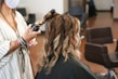 A person seated in a salon chair having their hair styled with a curling iron. The stylist is using the curling iron on a section of hair, resulting in soft, wavy curls. The atmosphere suggests a professional salon setting with additional styling chairs in the background.