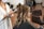 A person seated in a salon chair having their hair styled with a curling iron. The stylist is using the curling iron on a section of hair, resulting in soft, wavy curls. The atmosphere suggests a professional salon setting with additional styling chairs in the background.