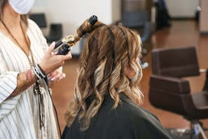 A person seated in a salon chair having their hair styled with a curling iron. The stylist is using the curling iron on a section of hair, resulting in soft, wavy curls. The atmosphere suggests a professional salon setting with additional styling chairs in the background.