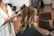 A person seated in a salon chair having their hair styled with a curling iron. The stylist is using the curling iron on a section of hair, resulting in soft, wavy curls. The atmosphere suggests a professional salon setting with additional styling chairs in the background.