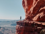 A person stands on the edge of a large red rock formation, taking a photograph. The landscape below is expansive, with distant mountains and a clear blue sky. The scale and vastness of the view suggest a sense of adventure and exploration.