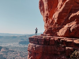A person stands on the edge of a large red rock formation, taking a photograph. The landscape below is expansive, with distant mountains and a clear blue sky. The scale and vastness of the view suggest a sense of adventure and exploration.