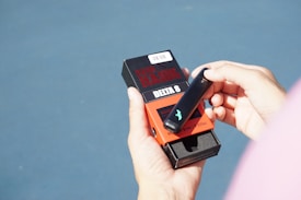 A person is holding a black vaping device and a dark-colored box labeled 'Dark Hawk Delta 8' with red accents. The person's hand is slightly tanned, and they are wearing a pink sleeve. The background is a smooth blue surface, possibly a table or a counter.