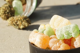 A collection of colorful gummy candies, mostly in the shapes of bears, are piled in a small black bowl. They appear to be coated in sugar. In the background, there are cannabis buds next to an overturned glass jar.