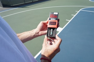 A person is holding a Dark Hawk Delta 8 product packaging in their hands. The background features a sports court with lines indicating the playing area. The person is wearing a white shirt, and the shot is taken in natural daylight.