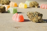 Colorful gummy candies and dried herbal buds are scattered on a flat surface. The focus is on a bright red gummy candy and a cannabis bud in the foreground, with other candies and buds slightly blurred in the background.