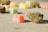 Colorful gummy candies and dried herbal buds are scattered on a flat surface. The focus is on a bright red gummy candy and a cannabis bud in the foreground, with other candies and buds slightly blurred in the background.