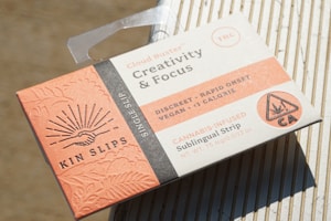 A packaged product labeled as Kin Slips, a cannabis-infused sublingual strip designed for creativity and focus. The packaging is rectangular with a predominantly peach and white color scheme, featuring a graphic of a handshake surrounded by rays of light. The package highlights key features such as being discreet, rapid onset, vegan, and having less than 1 calorie.
