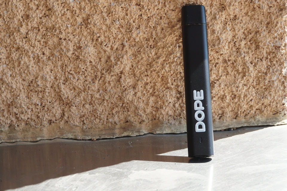 The image shows a black electronic device, likely an e-cigarette or vaporizer, standing upright on its side against a beige wall. The device is positioned slightly to the left and has a white label with the word "DOPE" written in bold black letters across it.