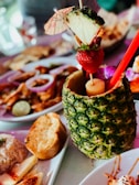 A vibrant tropical drink served in a hollowed-out pineapple, garnished with a skewer holding a slice of pineapple, a strawberry, and a cocktail umbrella. The foreground shows a variety of foods including rice, bread, and sliced fruits, suggesting a meal setting.