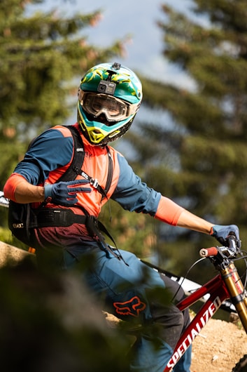 A mountain biker wearing a full-face helmet and protective gear is captured mid-trail ride. The rider is dressed in a blue and orange outfit, with a camera mounted on the helmet. The bike is red and has the word 'SPECIALIZED' on the frame. The background is blurred, indicating motion, with evergreen trees visible.