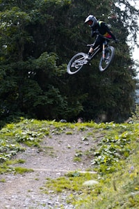 A person is performing an airborne stunt on a mountain bike, surrounded by lush green foliage and trees. The setting appears to be a dirt trail or off-road biking path, with greenery lining the track.