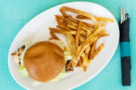 A grilled chicken sandwich topped with lettuce is placed on a round white plate alongside a portion of golden-brown French fries. The background is a textured turquoise tablecloth, and a fork and knife are wrapped in a black napkin with a turquoise band placed next to the plate.
