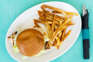 A grilled chicken sandwich topped with lettuce is placed on a round white plate alongside a portion of golden-brown French fries. The background is a textured turquoise tablecloth, and a fork and knife are wrapped in a black napkin with a turquoise band placed next to the plate.
