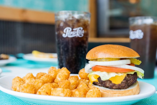 A cheeseburger topped with lettuce, tomato, onion, and pickles on a bun is served alongside a plate of crispy tater tots. In the background, two glasses of soda are on the table.
