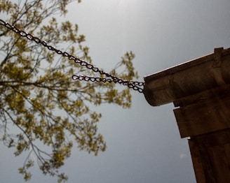 A metal gutter attached to a building with chains extends outward, with a tree branch in soft focus in the background against a blue sky. The sun casts a bright light, creating a serene and rustic atmosphere.