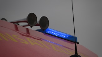 A close-up view of the top of an emergency vehicle featuring two large loudspeakers and a blue flashing light. The body of the vehicle is red with partially visible yellow lettering.