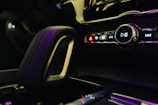 Close-up of a car's interior focusing on the multimedia control panel and gear shift. The panel includes buttons with symbols for play, pause, skip, and hazard lights. The lighting is dim with a soft glow from pink and purple hues reflecting off the surfaces.