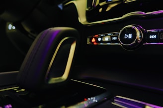 Close-up of a car's interior focusing on the multimedia control panel and gear shift. The panel includes buttons with symbols for play, pause, skip, and hazard lights. The lighting is dim with a soft glow from pink and purple hues reflecting off the surfaces.