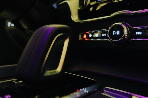 Close-up of a car's interior focusing on the multimedia control panel and gear shift. The panel includes buttons with symbols for play, pause, skip, and hazard lights. The lighting is dim with a soft glow from pink and purple hues reflecting off the surfaces.