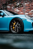 A vibrant blue sports car with a focus on its sleek body and prominent black alloy wheel with yellow brake calipers, set against a textured brick wall in an urban environment.