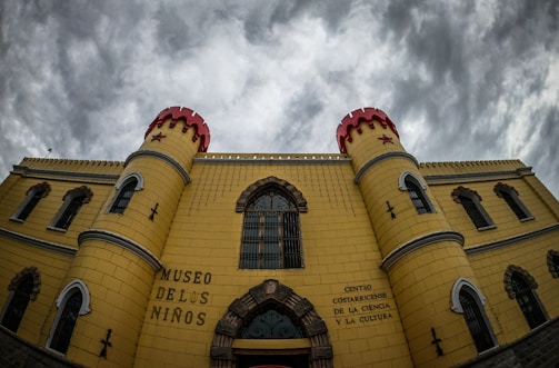 A castle-like building with yellow walls and red turret tops stands under a cloudy sky. The facade includes multiple arched windows with bars, and the entrance is highlighted by a large arched doorway. The walls have inscriptions in Spanish, identifying the building as the 'Museo de los Niños' and the 'Centro Costarricense de la Ciencia y la Cultura'.