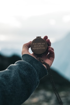 A hand is holding a brass compass on which the words 'YOU ARE MY FAVORITE ADVENTURE' are engraved. The background shows an out-of-focus landscape with a bright sky, suggesting a sense of travel or exploration.
