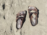 A pair of brown, open-toed sandals with straps and buckles are lying on a sandy surface. The sand appears to be beach sand, and the sandals seem to be well-worn.