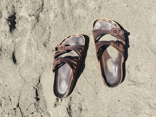 A pair of brown, open-toed sandals with straps and buckles are lying on a sandy surface. The sand appears to be beach sand, and the sandals seem to be well-worn.