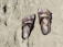 A pair of brown, open-toed sandals with straps and buckles are lying on a sandy surface. The sand appears to be beach sand, and the sandals seem to be well-worn.