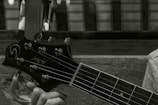 A close-up image of a person's hand holding the neck and headstock of a guitar. The guitar headstock has tuning pegs and a logo, with strings running down the neck. The background appears to be an out-of-focus window or wall.